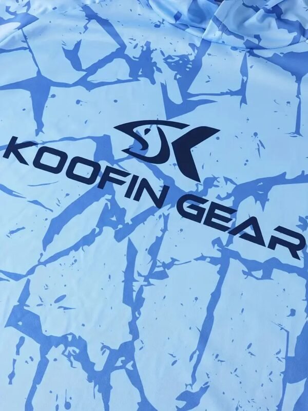 هذه صورة مقربة لتيشيرت صيد يحمل شعار "KOOFIN GEAR" بتصميم أزرق مموه، يظهر تفاصيل النقوش والنسيج.